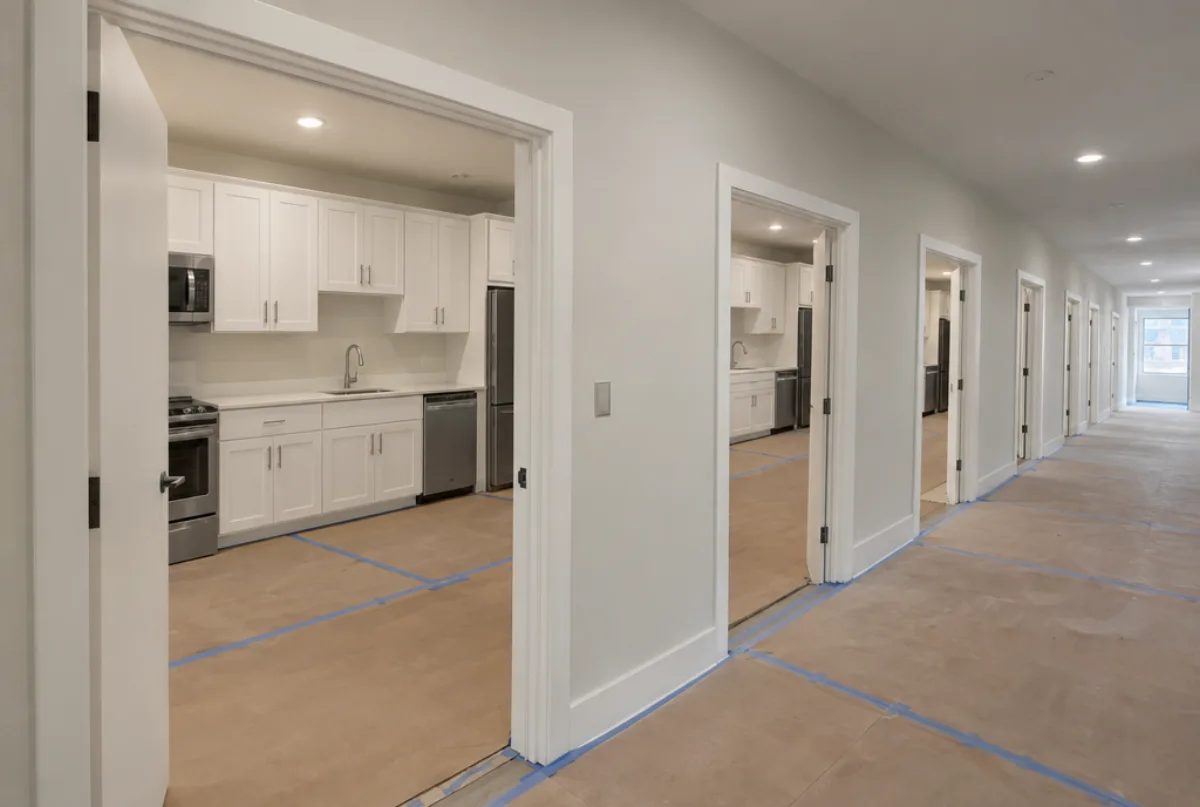 Corridor of a new apartment building with open doors showing identical white shaker kitchens, floors covered in protective paper